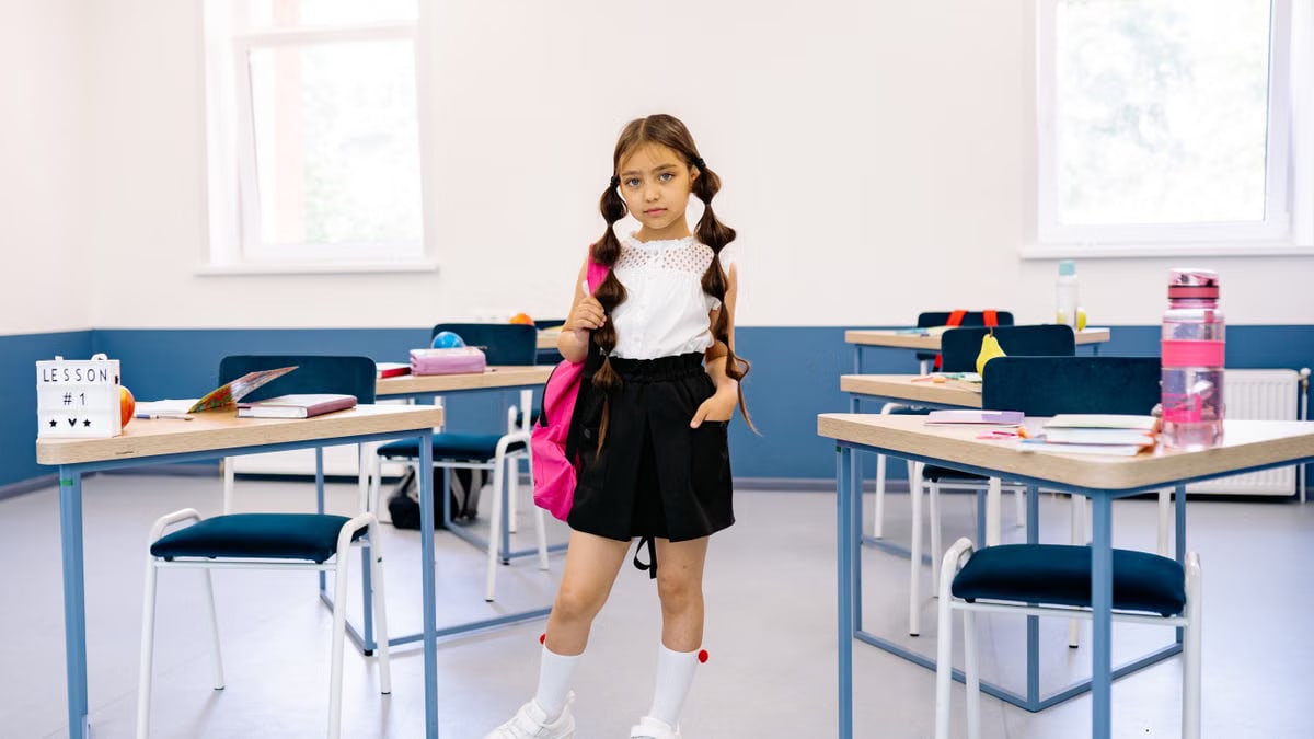 A young girl stands confidently in a bright, modern classroom with desks and educational materials.